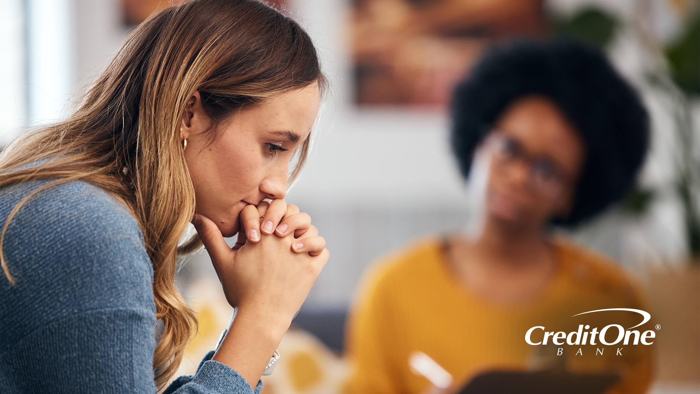 A young woman rests her chin on her hands as if she’s contemplating a crisis while another woman, who is perhaps her therapist or credit counselor, looks on with a clipboard.